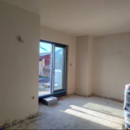 Wnętrze w trakcie prac wykończeniowych, ściany po nałożeniu tynku maszynowego, widoczne puszki elektryczne, worki z zaprawą Knauf MP 75 Diamant oraz wyjście na zewnątrz z widokiem na budynek...