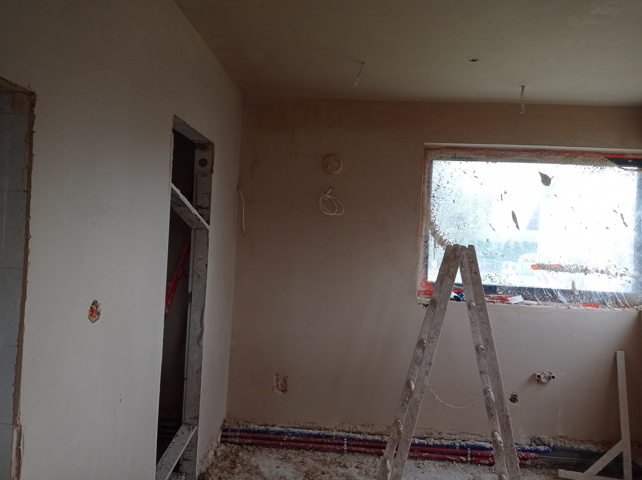 Wnętrze w trakcie prac wykończeniowych z nałożonymi tynkami: widoczne ściany pokryte świeżym tynkiem, drabina oparta o okno zabezpieczone folią, instalacje podłogowe oraz otwory pod gniazdka...