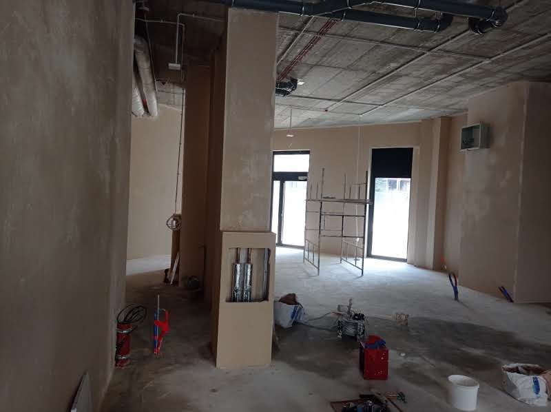 Wnętrze budynku w trakcie prac wykończeniowych, świeżo nałożone tynki na ścianach i filarach, widoczne instalacje w otworach, rusztowanie, sprzęt i narzędzia do tynkowania rozstawione na podłodze.