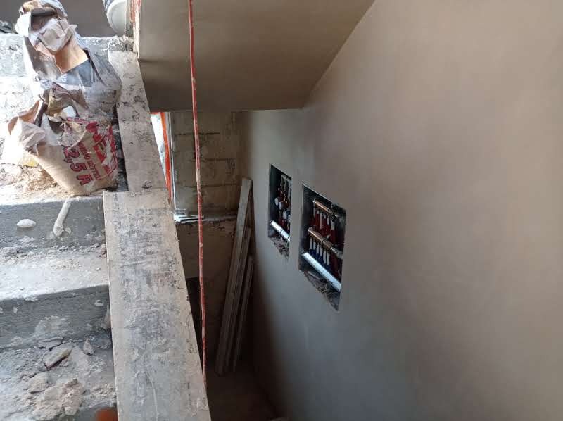 Świeżo nałożony, gładki tynk na ścianie wewnętrznej, z widocznymi otworami na instalacje, obok schody z workiem zaprawy i deskami.