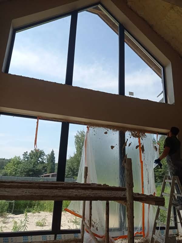 Prace tynkarskie w nowym domu z dużymi oknami, zabezpieczonymi folią, widok na zewnątrz z rusztowania, pracownik w tle.