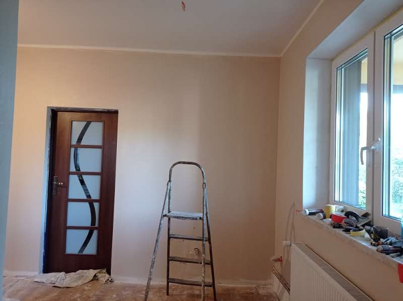 Pokój po nałożeniu gładzi, widoczna drabina malarska, okno z narzędziami i akcesoriami malarskimi, drzwi z dekoracyjnym szkłem, ściany w odcieniach beżu.