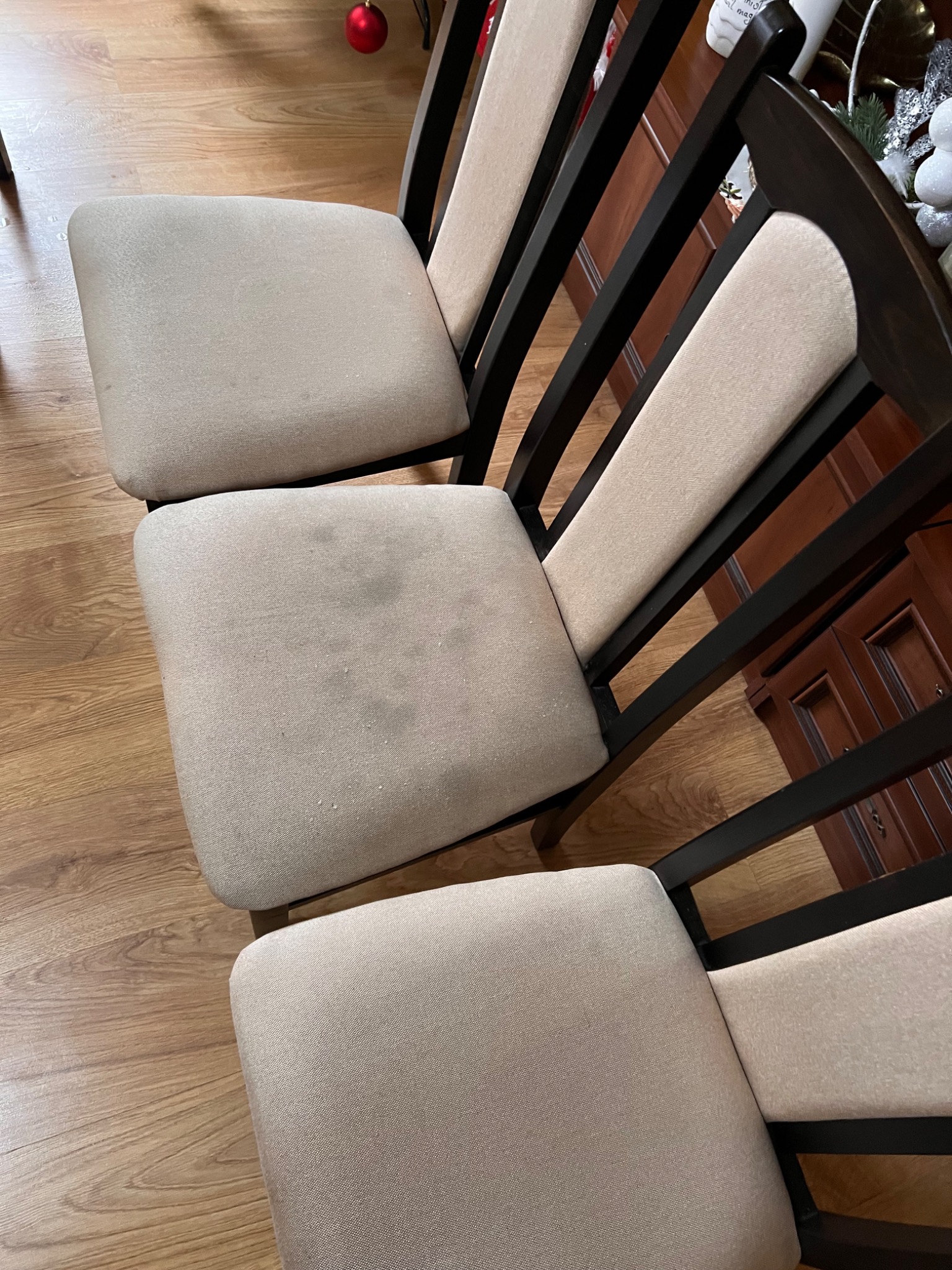 Trzy krzesła z beżową tapicerką i ciemnobrązowymi ramami, widoczne plamy na środkowym siedzisku, na tle drewniana podłoga i fragment komody.