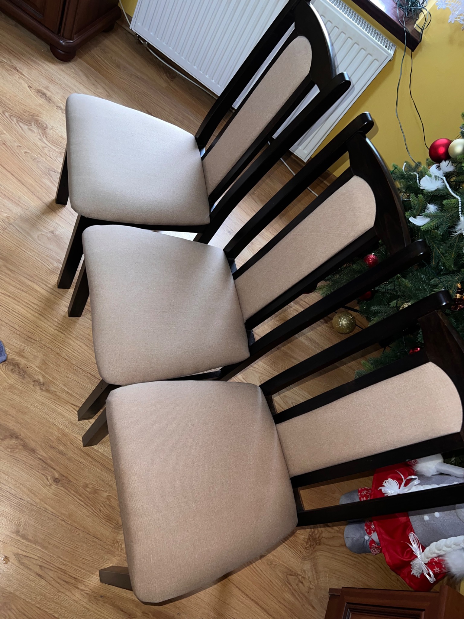 Trzy krzesła z beżową tapicerką i ciemnobrązową ramą ustawione jeden na drugim, widoczne na tle drewnianej podłogi, grzejnika i choinki z ozdobami.