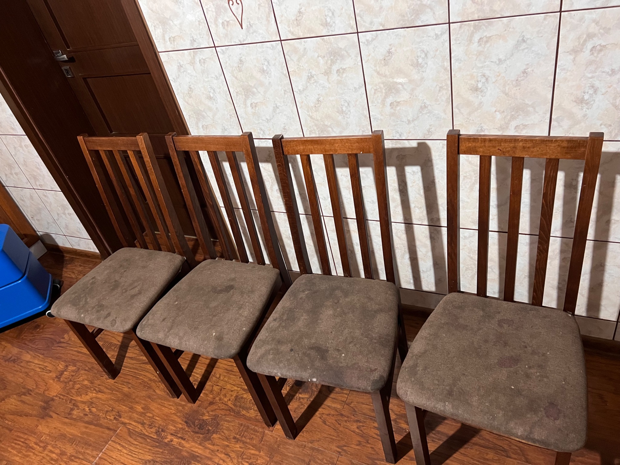Cztery drewniane krzesła z brązowymi, miejscami zabrudzonymi siedziskami, ustawione rzędem na parkiecie, z fragmentem niebieskiego odkurzacza widocznym z lewej strony.