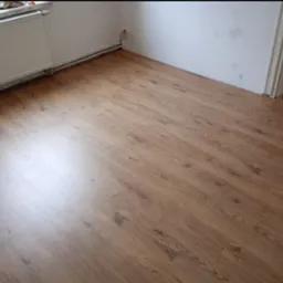 Ułożone panele podłogowe w odcieniach brązu, widoczny grzejnik i wystające kable ze ściany w rogu pomieszczenia.