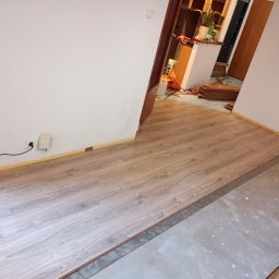 Piotr Szkiel - W trakcie układania paneli podłogowych w pomieszczeniu, widoczne panele o wzorze drewna na części podłogi, reszta powierzchni przygotowana, zabezpieczona taśmą malarską przy ścianach, w tle sterta...