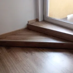 Dwa stopnie schodów wewnętrznych wykończone panelami drewnopodobnymi, z widoczną listwą przypodłogową i fragmentem podłogi, przy oknie z widokiem na zewnętrzne schody.