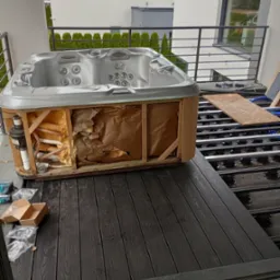 Montaż wanny spa na balkonie: widoczna konstrukcja wanny z instalacją, częściowo usunięta izolacja, czarne deski tarasowe i elementy konstrukcyjne pod spodem.