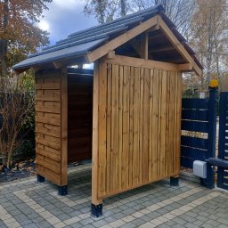 Piotr Szkiel - Drewniana wiata z szarym dachem na kostce brukowej, widoczna z boku, obok brama wjazdowa z automatyką. Jesienne drzewa w tle.