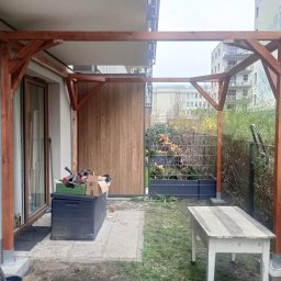 Piotr Szkiel - Drewniana pergola w trakcie budowy, widoczne narzędzia i materiały. Tło stanowi budynek mieszkalny i zieleń. Ujęcie z bliska, ukazujące detale konstrukcji.