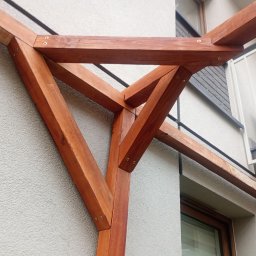 Piotr Szkiel - Fragment drewnianej pergoli z widocznymi połączeniami śrubowymi, na tle elewacji budynku i balkonu z białą balustradą. Ujęcie z dołu, podkreślające konstrukcję.