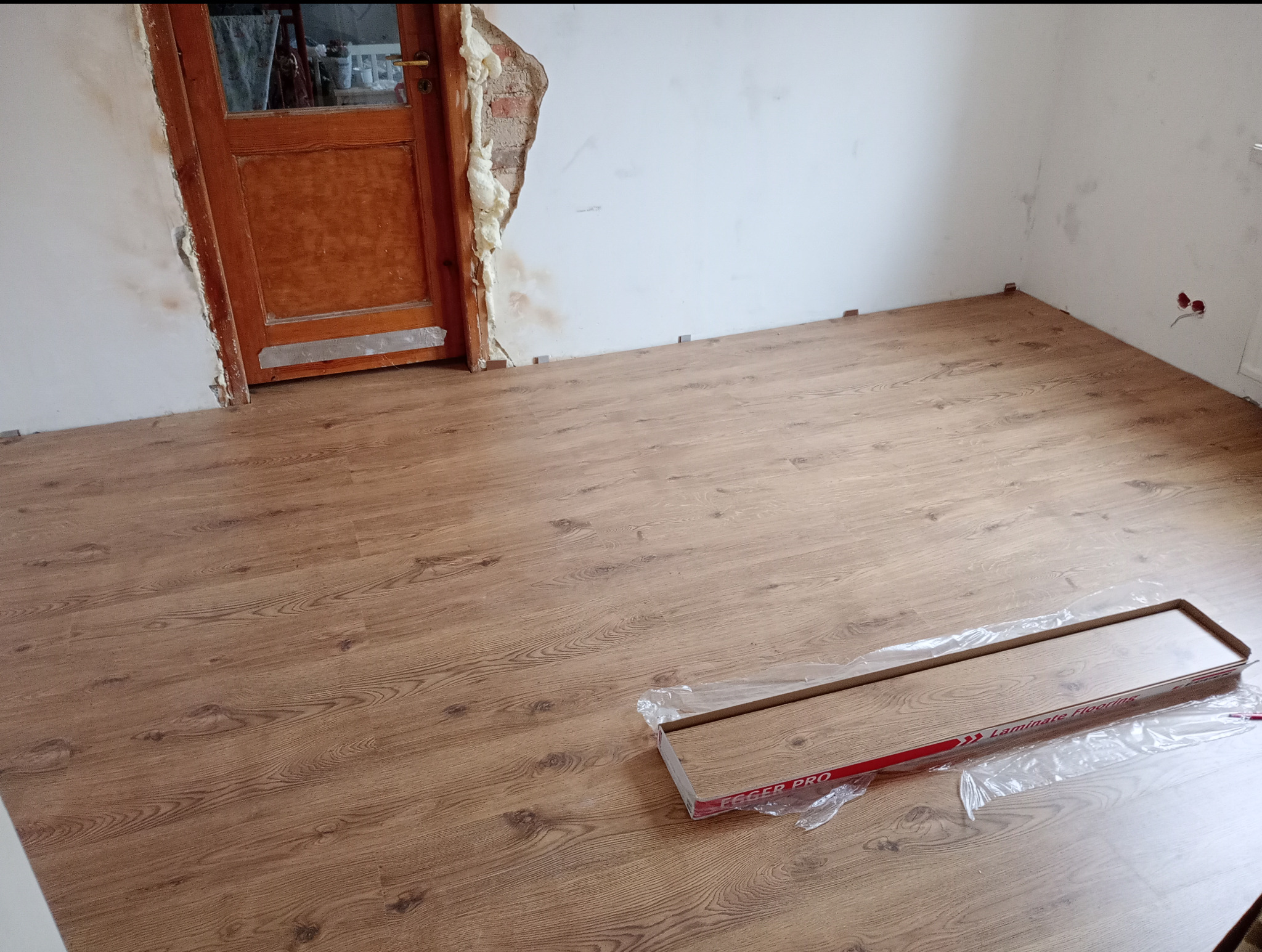 Układanie paneli podłogowych w trakcie remontu, widoczna paczka paneli 'Eager Pro Laminate Flooring' w folii, fragment ściany z odkrytą cegłą i pianką montażową przy drewnianych drzwiach.