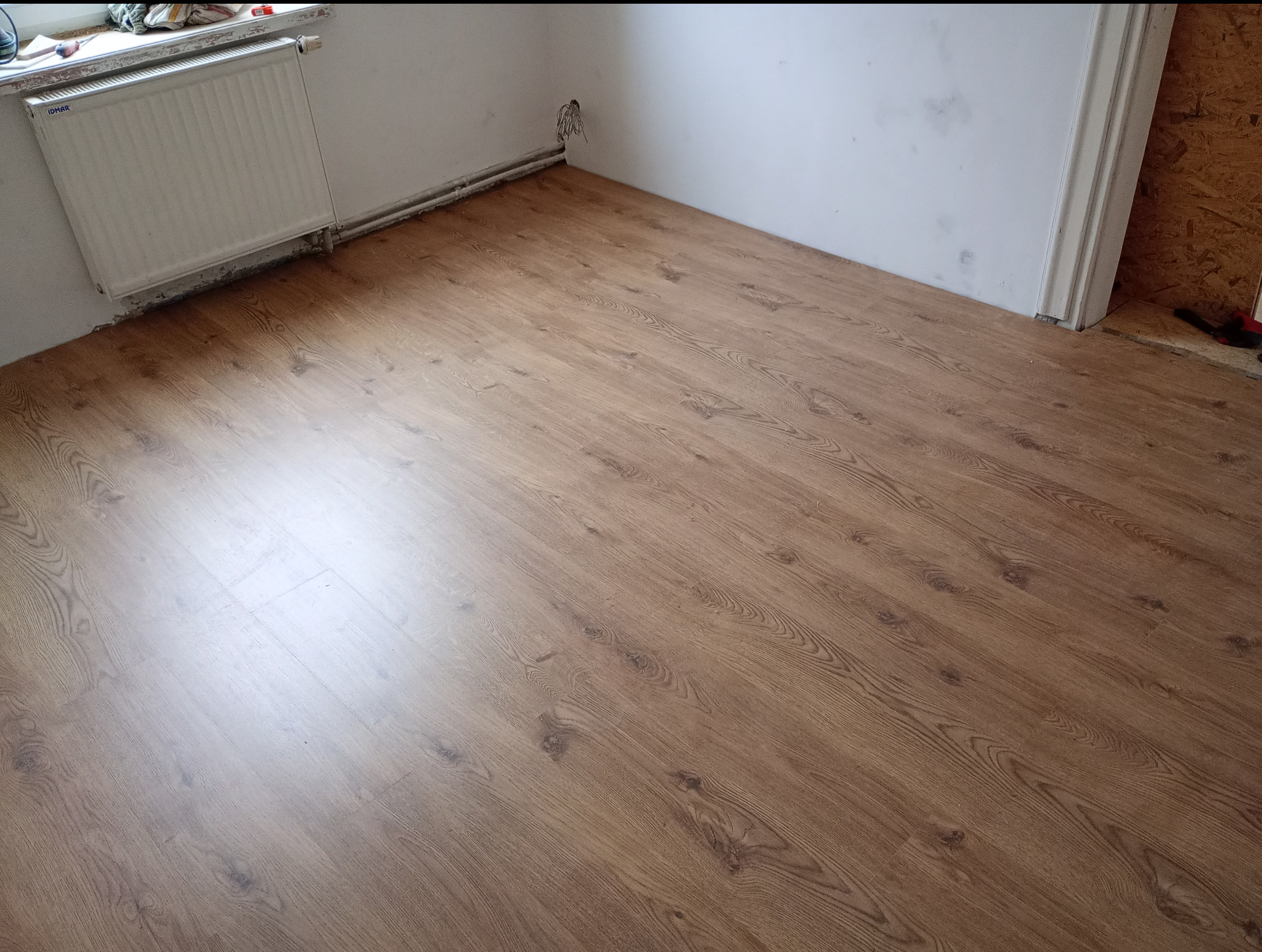 Ułożone panele podłogowe w odcieniach brązu, widoczny grzejnik i wystające kable ze ściany w rogu pomieszczenia.