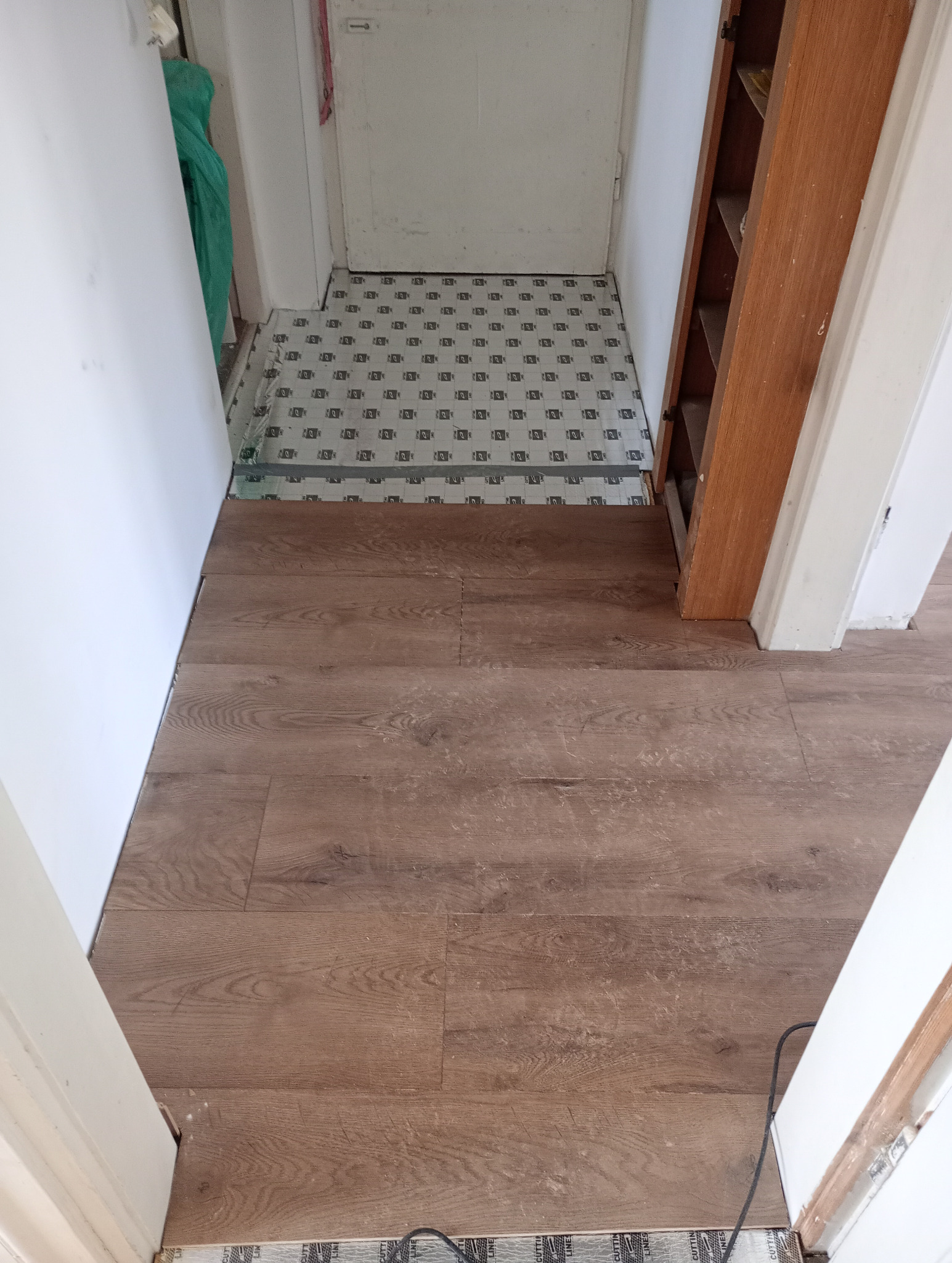 Fragment korytarza z częściowo ułożoną podłogą z paneli imitujących drewno. Widoczna podkładka pod panele z nadrukiem. W tle drzwi i drewniany regał.