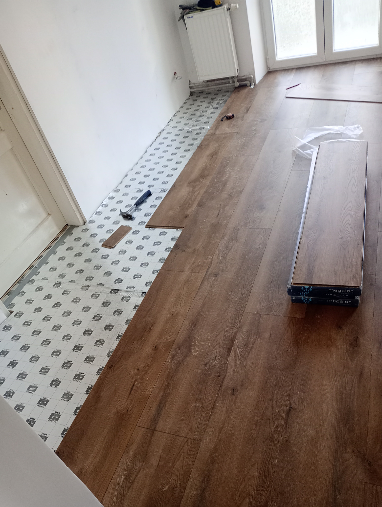 Układanie paneli podłogowych w trakcie pracy: widoczne panele Megaloc, podkład, młotek i częściowo ułożona podłoga z paneli drewnopodobnych.