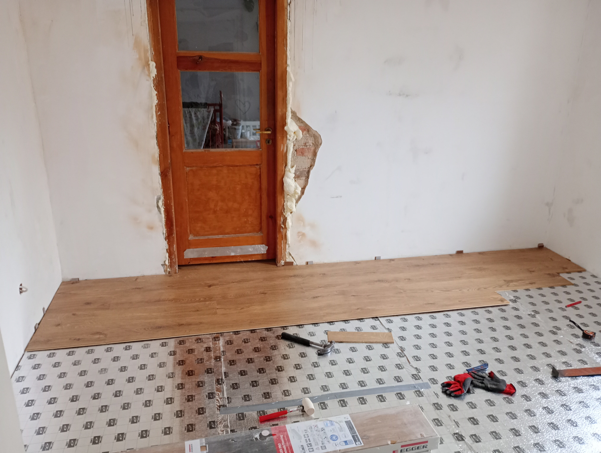 Proces układania paneli podłogowych w pomieszczeniu z niedokończonymi ścianami, widoczne narzędzia, podkład izolacyjny, panele Egger i szczelina wokół ościeżnicy drzwiowej wypełniona pianką montażową.