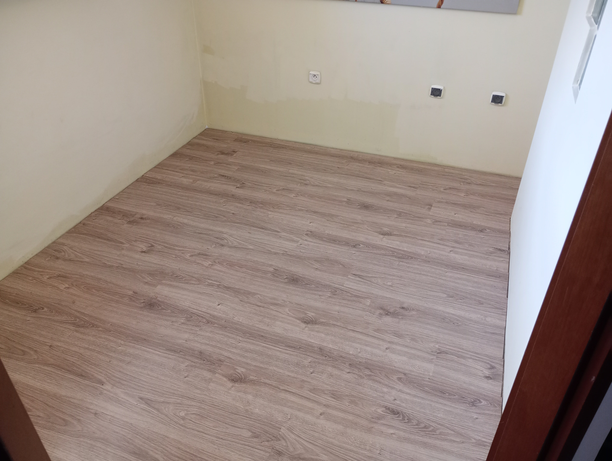 Ułożone panele podłogowe o wzorze drewna w pustym pokoju z pomalowanymi ścianami i widocznymi gniazdkami elektrycznymi.