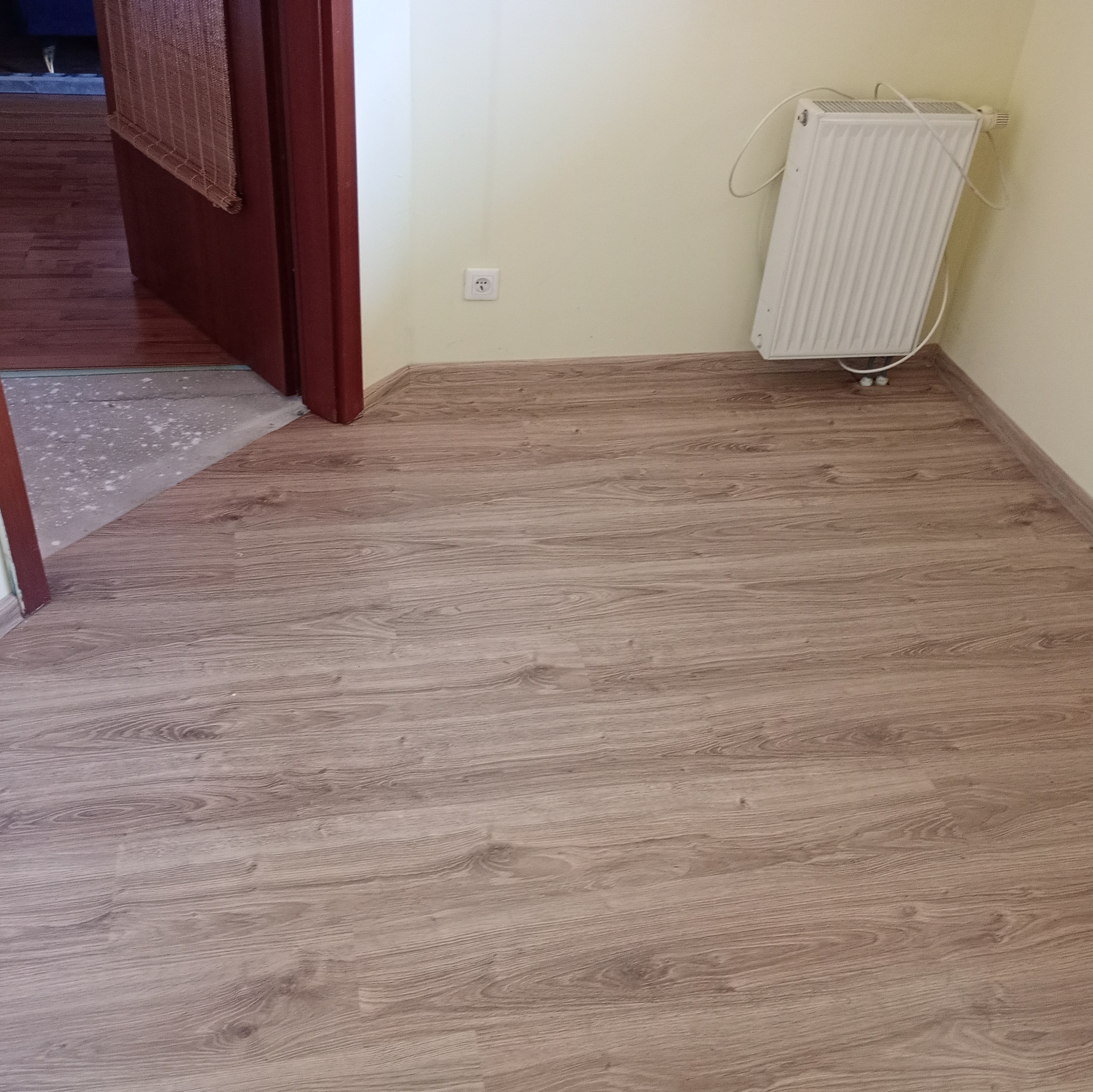 Ułożone panele podłogowe w pomieszczeniu z grzejnikiem i otwartymi drzwiami, widoczny fragment wylewki.