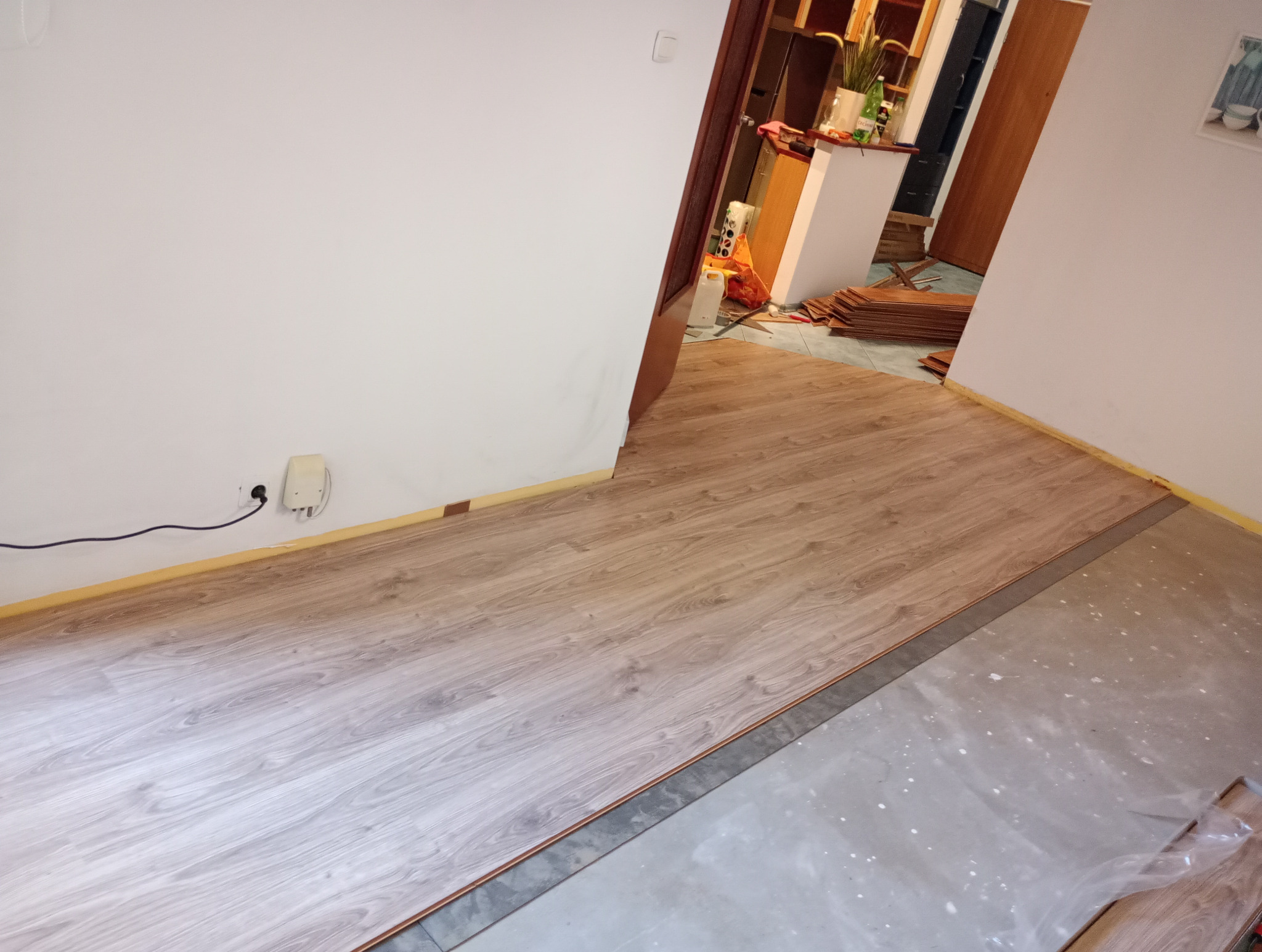 W trakcie układania paneli podłogowych w pomieszczeniu, widoczne panele o wzorze drewna na części podłogi, reszta powierzchni przygotowana, zabezpieczona taśmą malarską przy ścianach, w tle sterta...