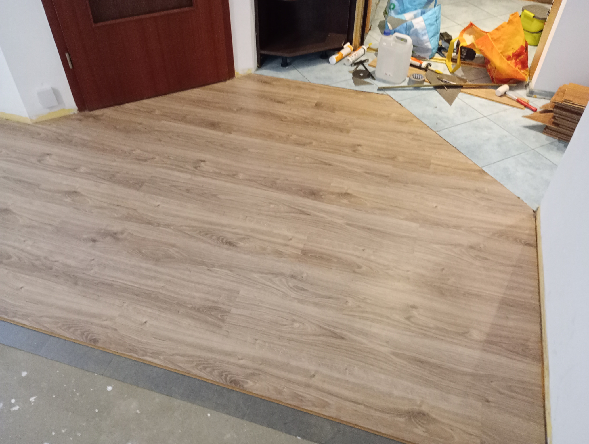 Fragment układanej podłogi z paneli w kształcie zbliżonym do trójkąta, z widocznymi narzędziami i materiałami budowlanymi w tle, w tym biały pojemnik, tuba z klejem oraz pomarańczowa torba.