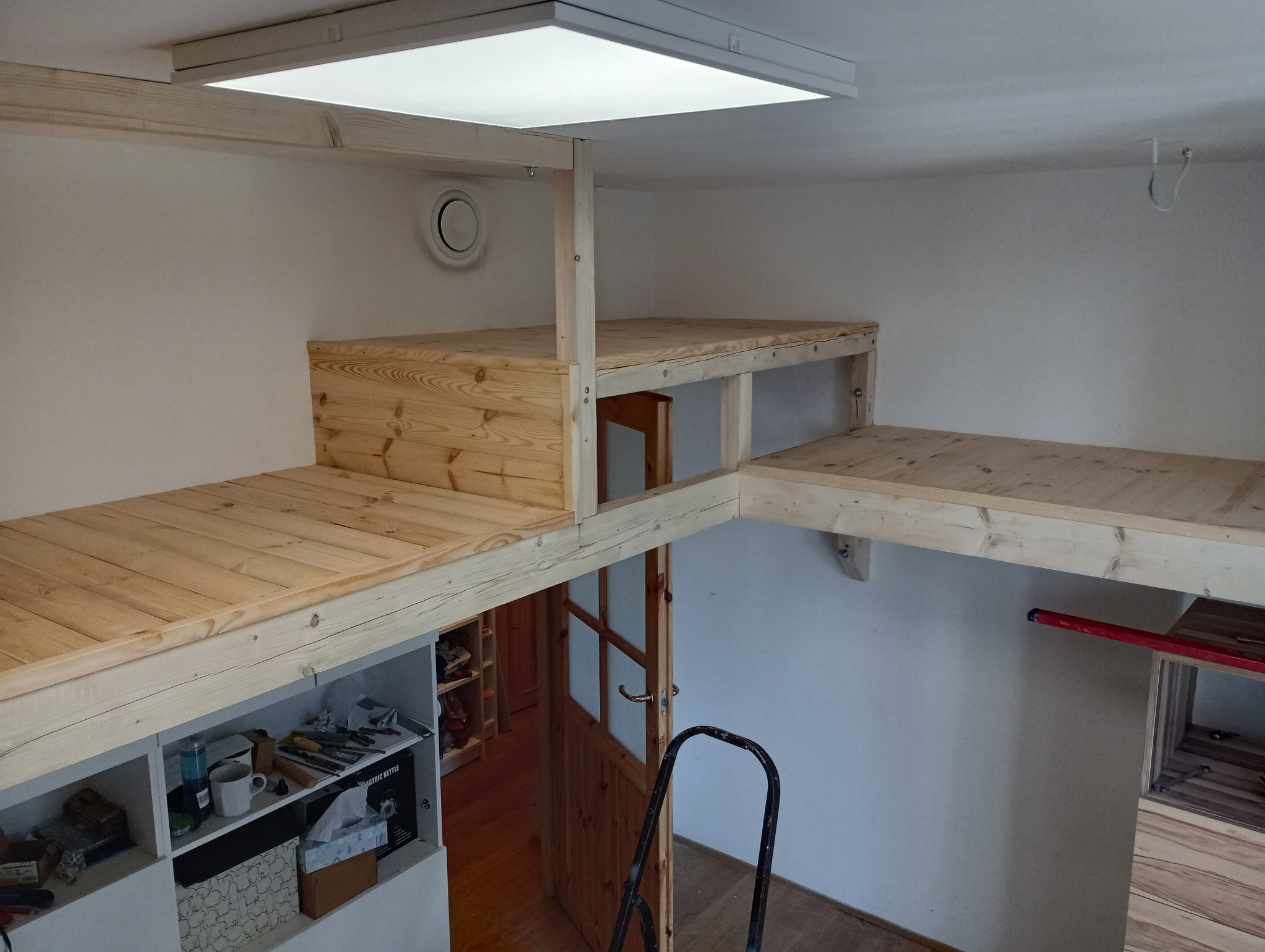 Wielopoziomowa konstrukcja z jasnego drewna w pokoju, z drabinką opartą o drzwi, widoczna szafka z półkami pod spodem.