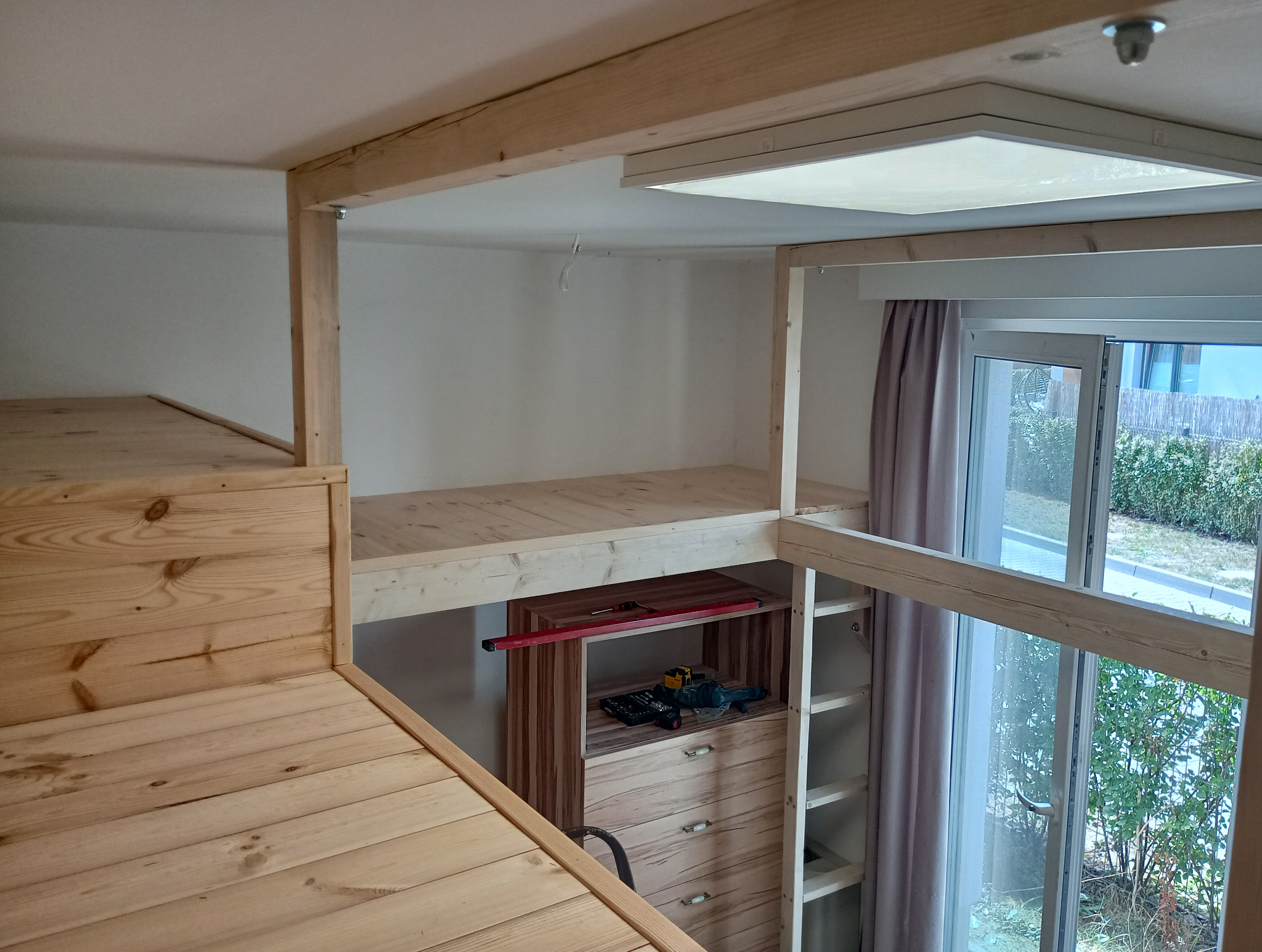 Drewniana konstrukcja antresoli z widocznymi schodami, półkami i narzędziami, częściowo oświetlona przez okno balkonowe.