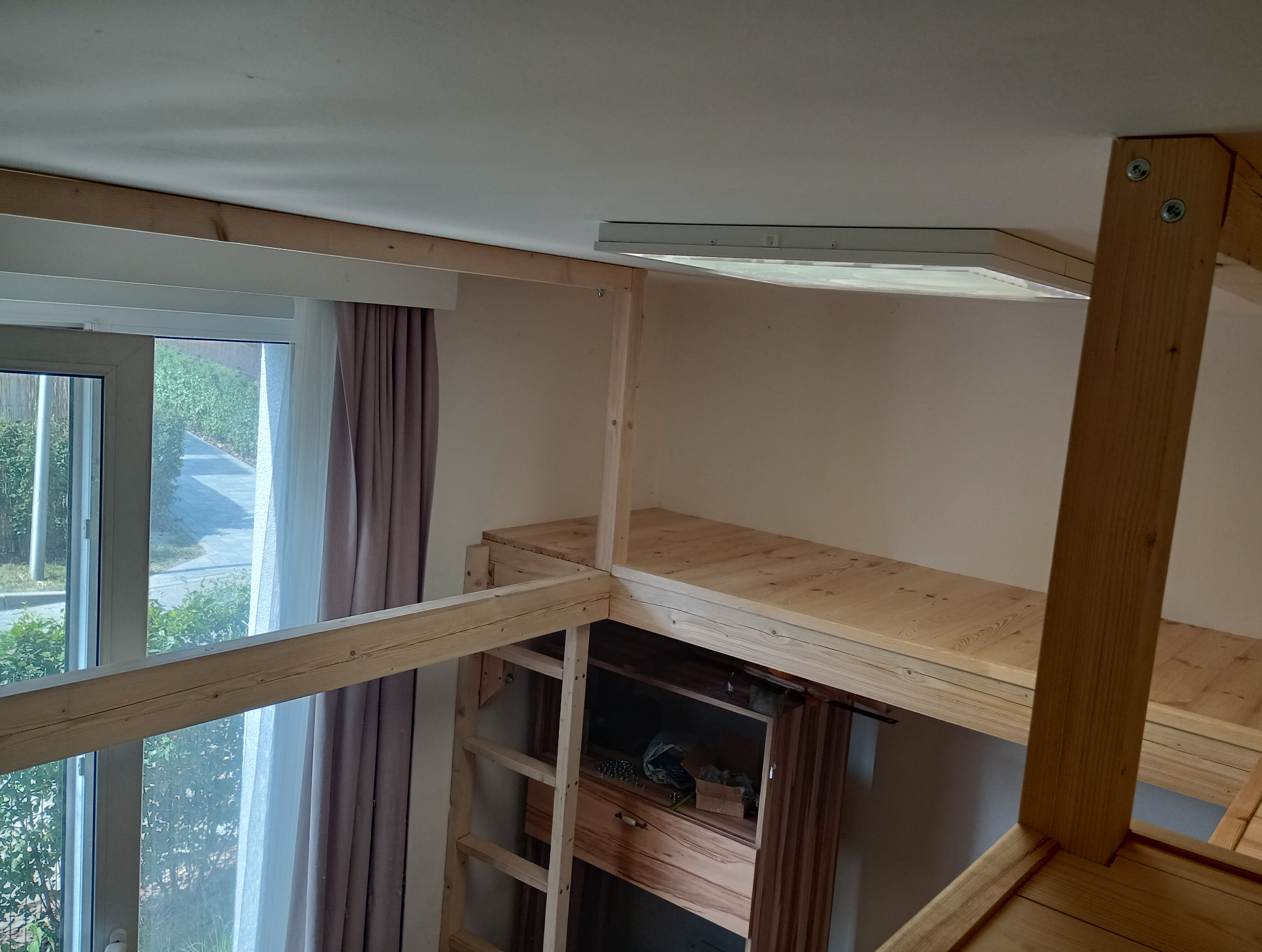 Drewniana konstrukcja antresoli z drabinką w jasnym wnętrzu, widoczne elementy mocujące i fragment okna z zasłonami.