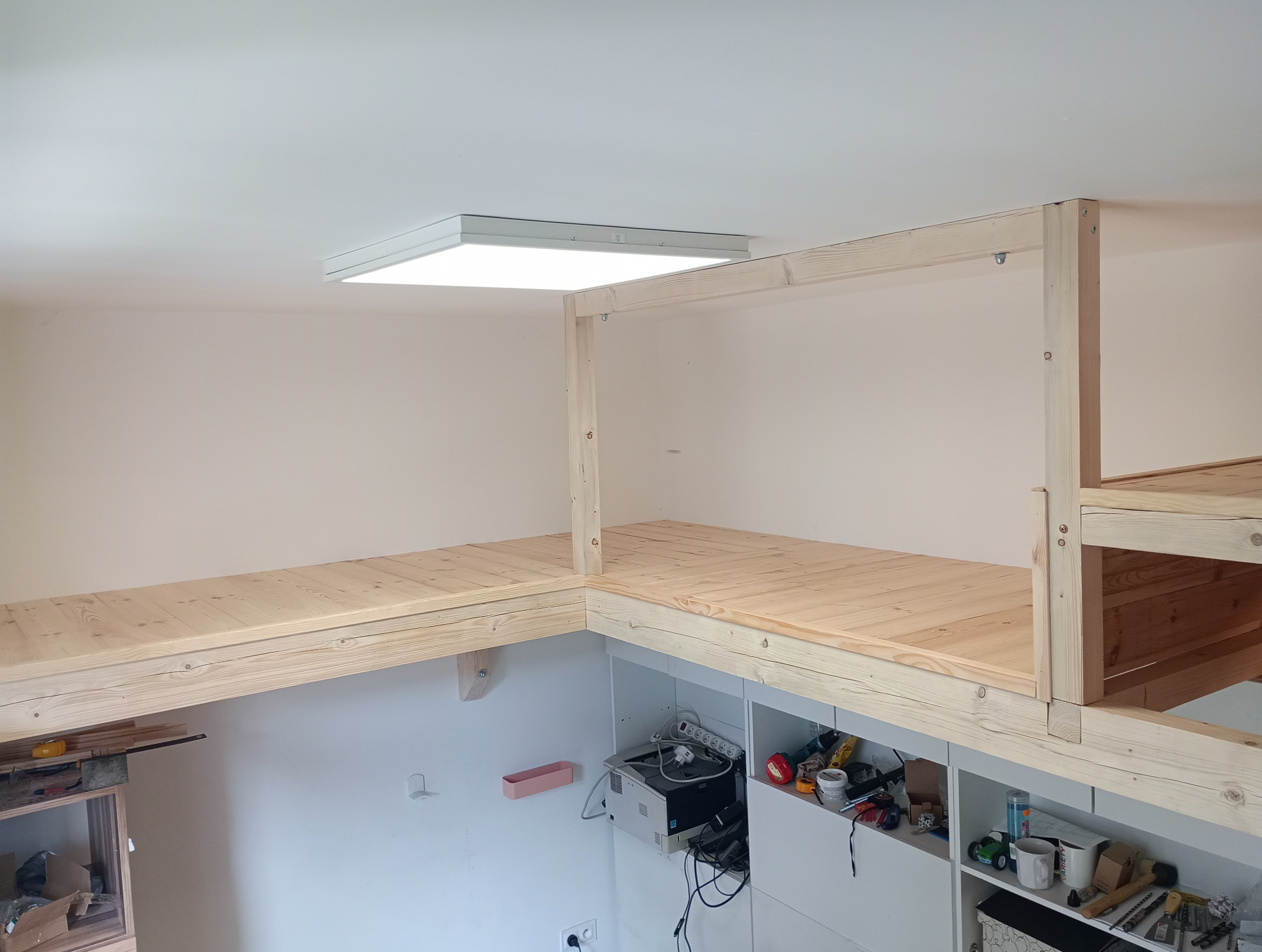 Drewniana antresola z jasnego drewna w pokoju, widok z dołu na konstrukcję i sufit z oświetleniem LED, widoczne narzędzia i przedmioty pod antresolą.