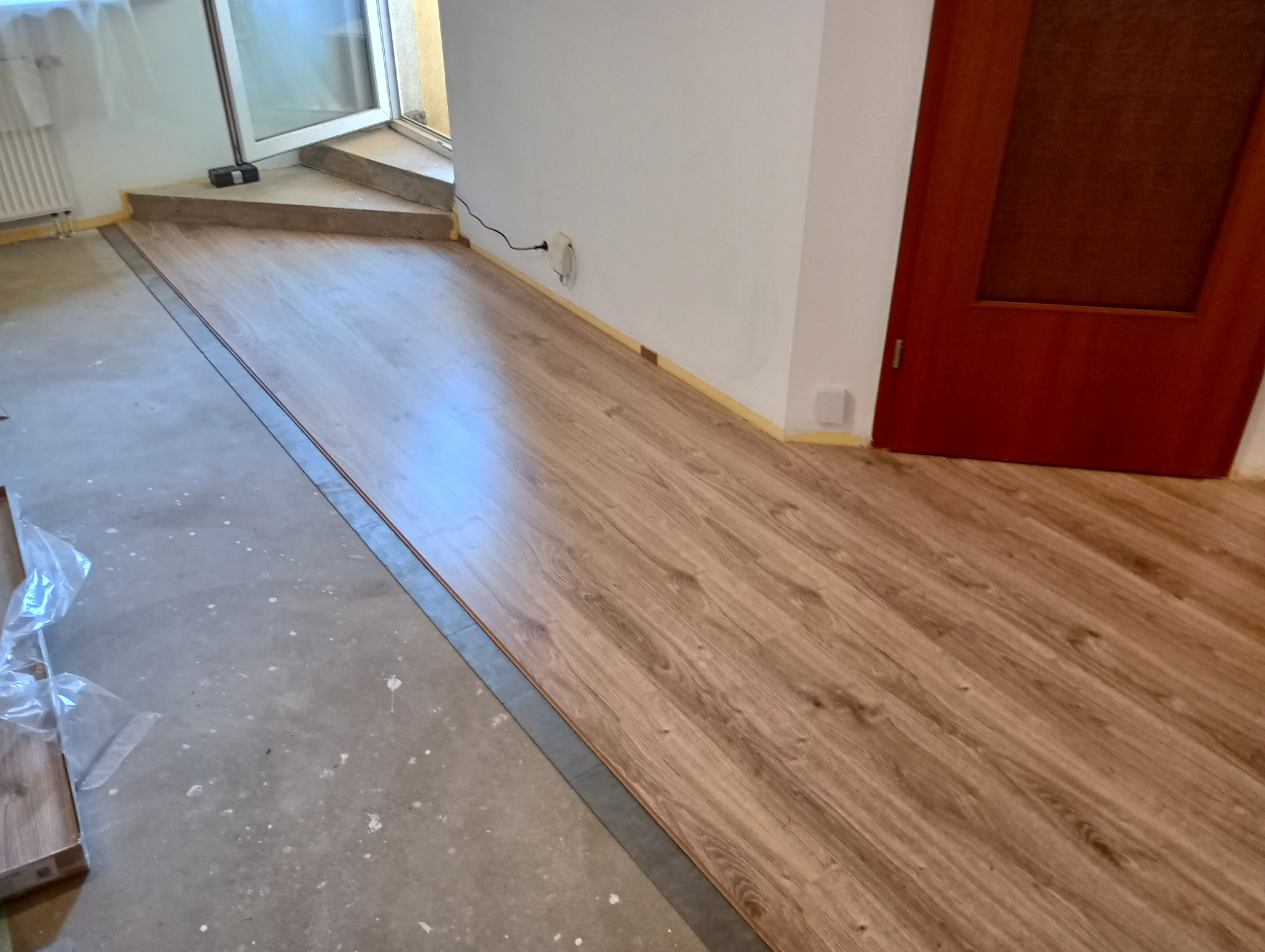 Układanie paneli podłogowych o wzorze drewna w trakcie montażu, widoczny fragment surowej podłogi i przygotowane panele w folii.