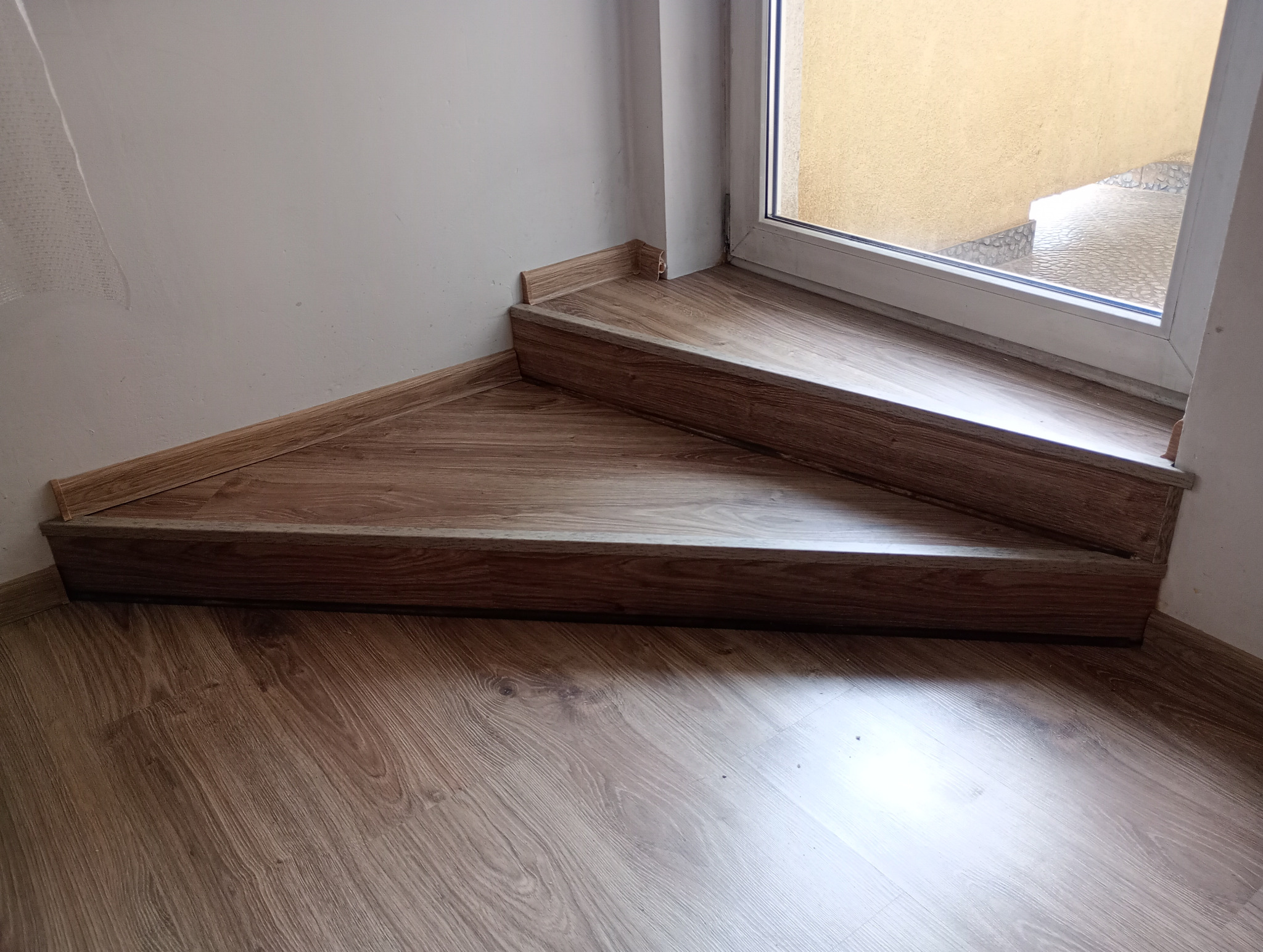 Dwa stopnie schodów wewnętrznych wykończone panelami drewnopodobnymi, z widoczną listwą przypodłogową i fragmentem podłogi, przy oknie z widokiem na zewnętrzne schody.