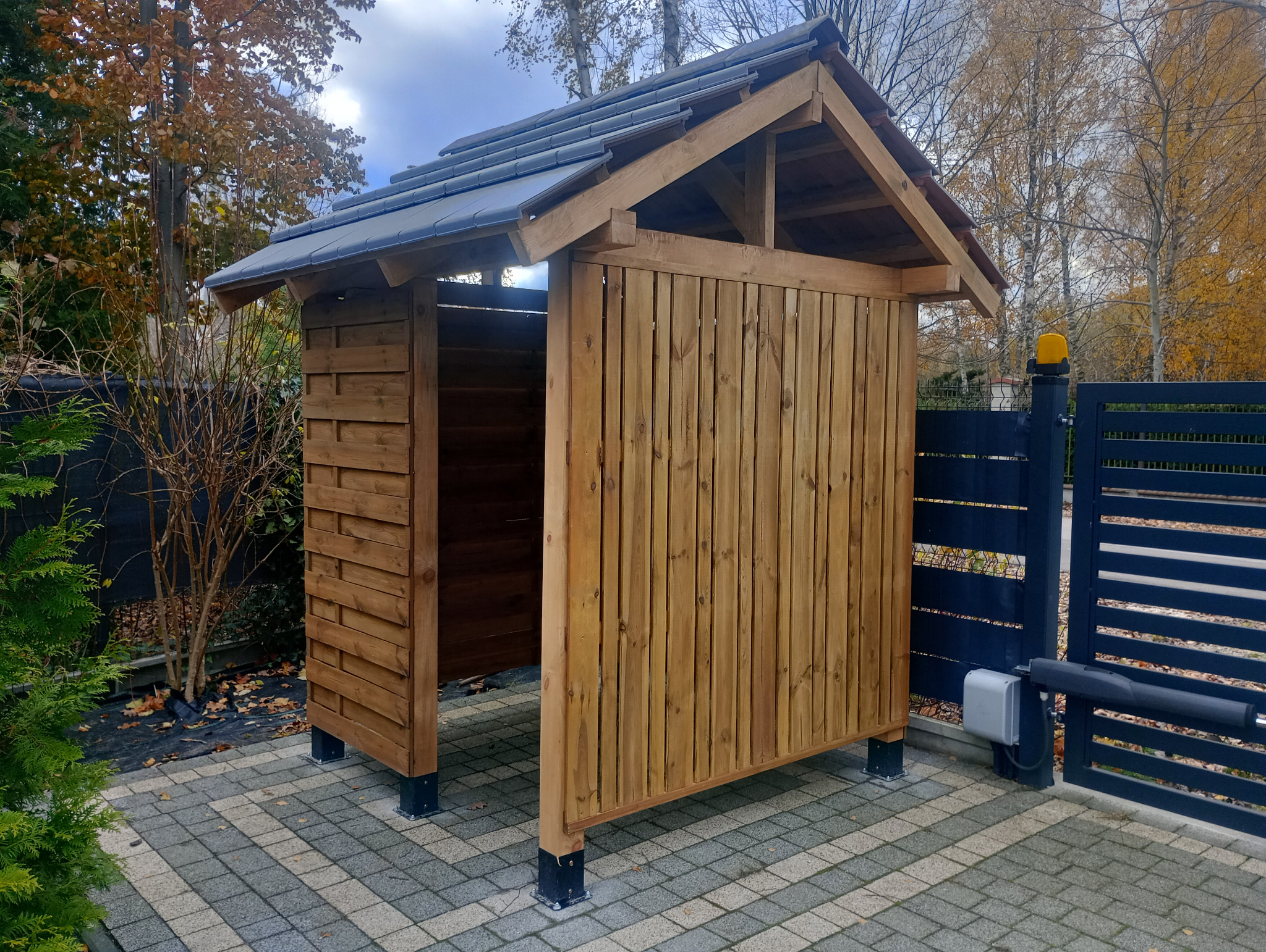 Drewniana wiata z szarym dachem na kostce brukowej, widoczna z boku, obok brama wjazdowa z automatyką. Jesienne drzewa w tle.
