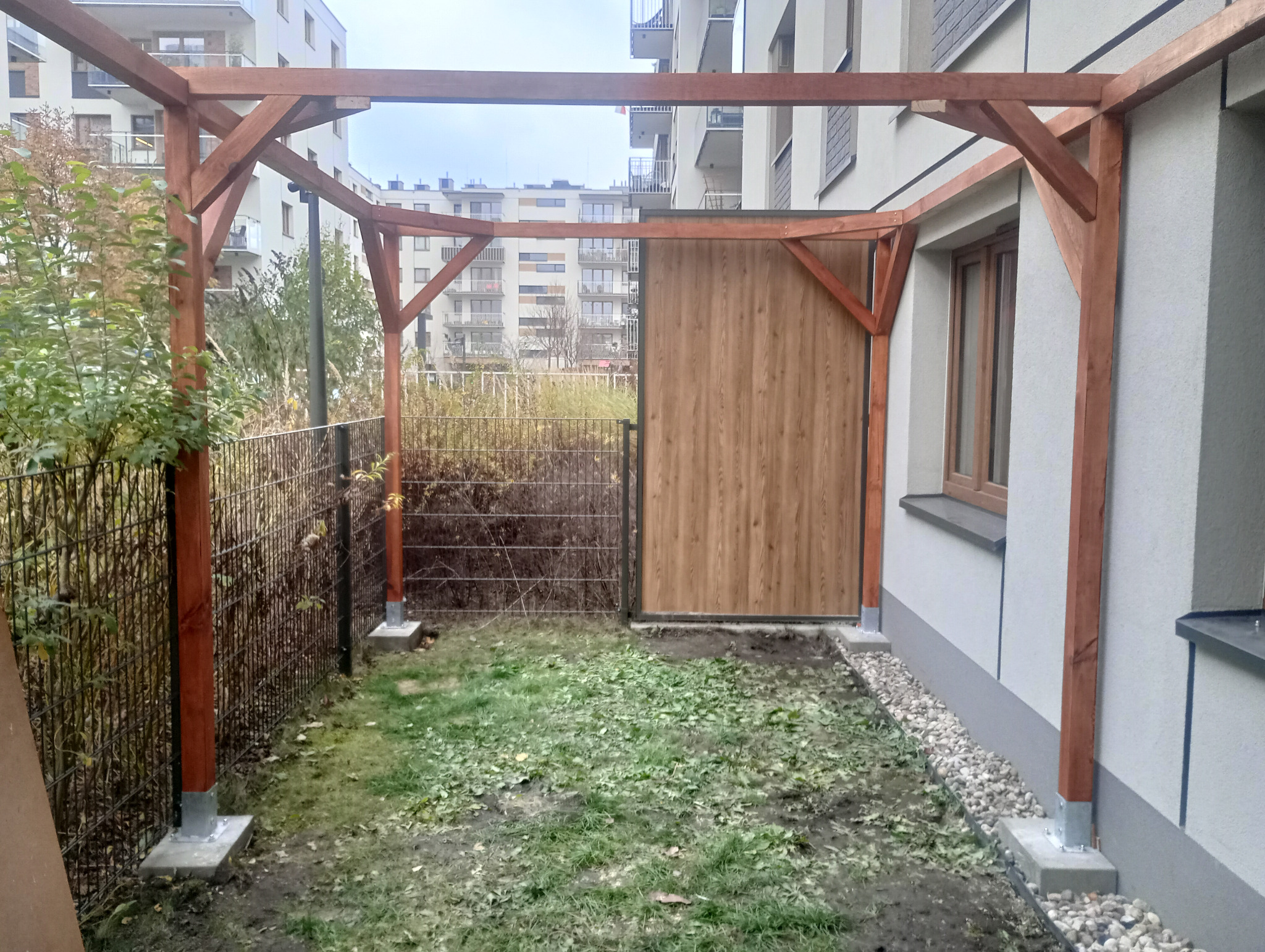 Drewniana pergola z zadaszeniem częściowo osłoniętym panelami, widok z perspektywy ogrodu, na tle zabudowy wielorodzinnej i ogrodzenia.