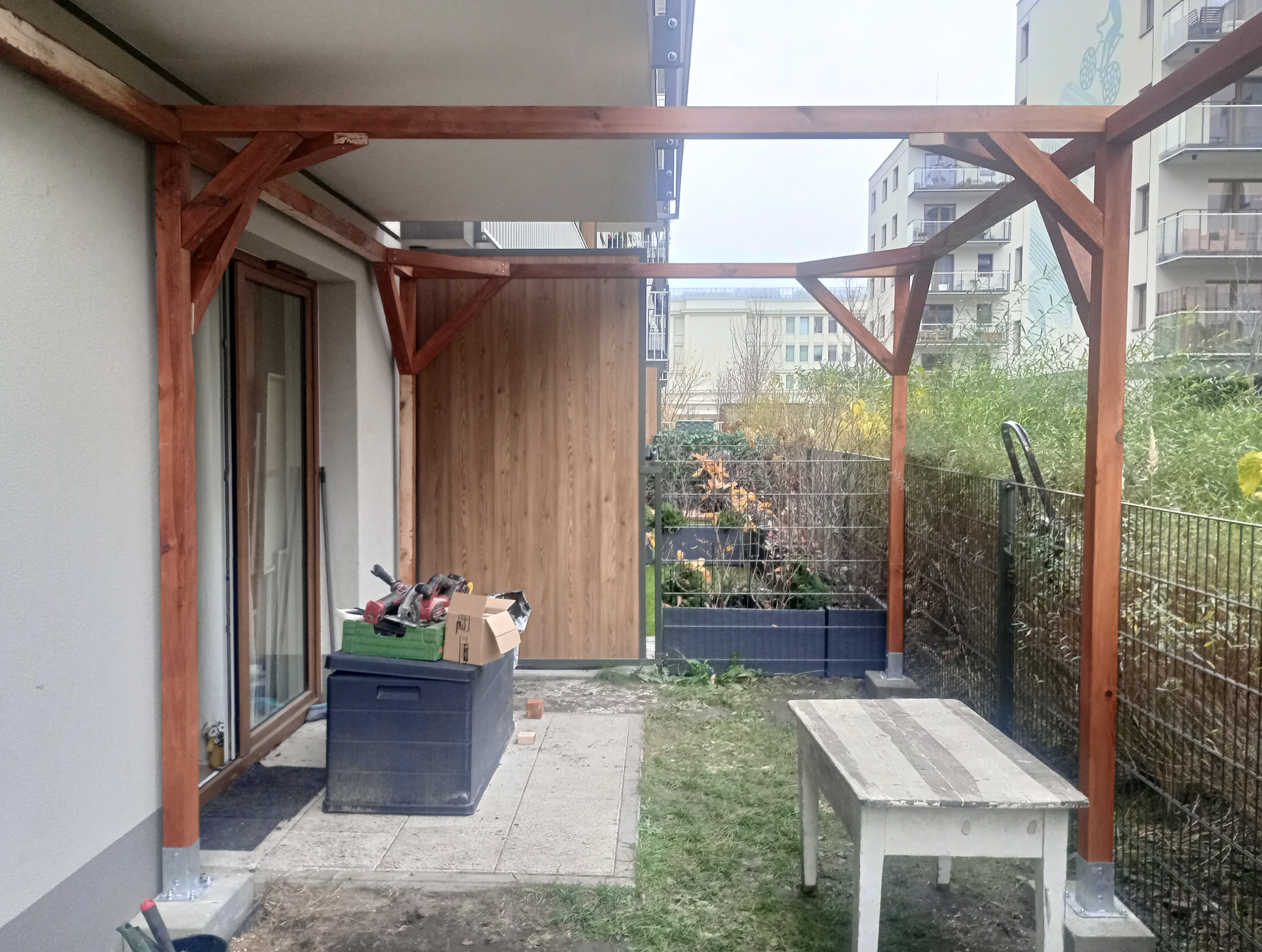 Drewniana pergola w trakcie budowy, widoczne narzędzia i materiały. Tło stanowi budynek mieszkalny i zieleń. Ujęcie z bliska, ukazujące detale konstrukcji.