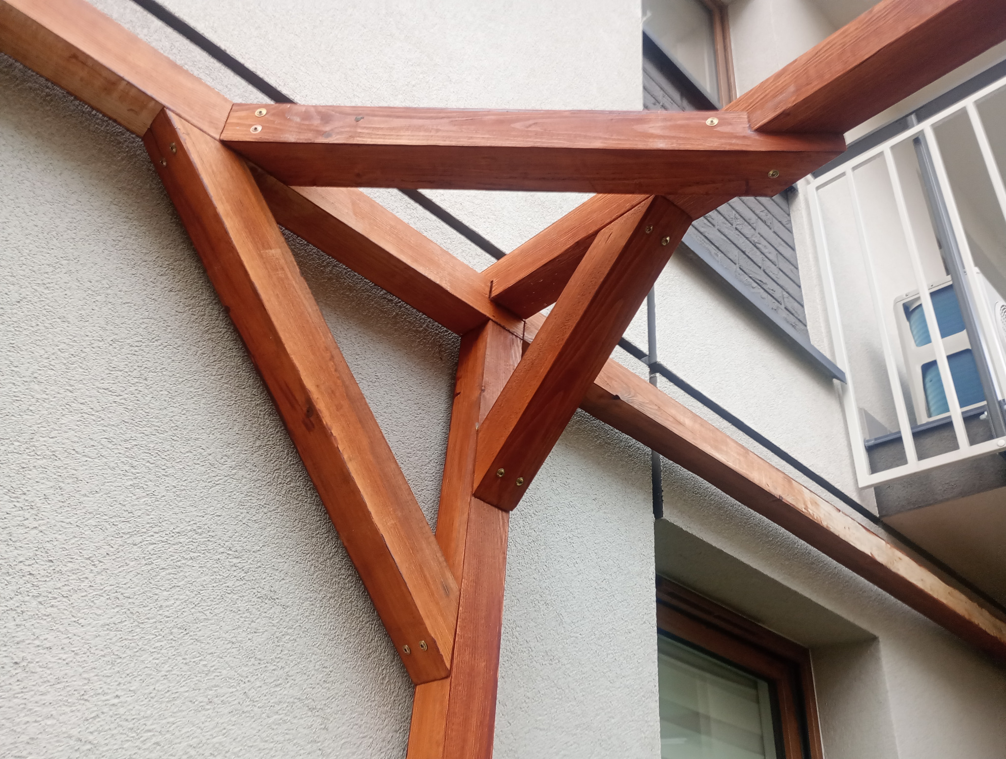 Fragment drewnianej pergoli z widocznymi połączeniami śrubowymi, na tle elewacji budynku i balkonu z białą balustradą. Ujęcie z dołu, podkreślające konstrukcję.