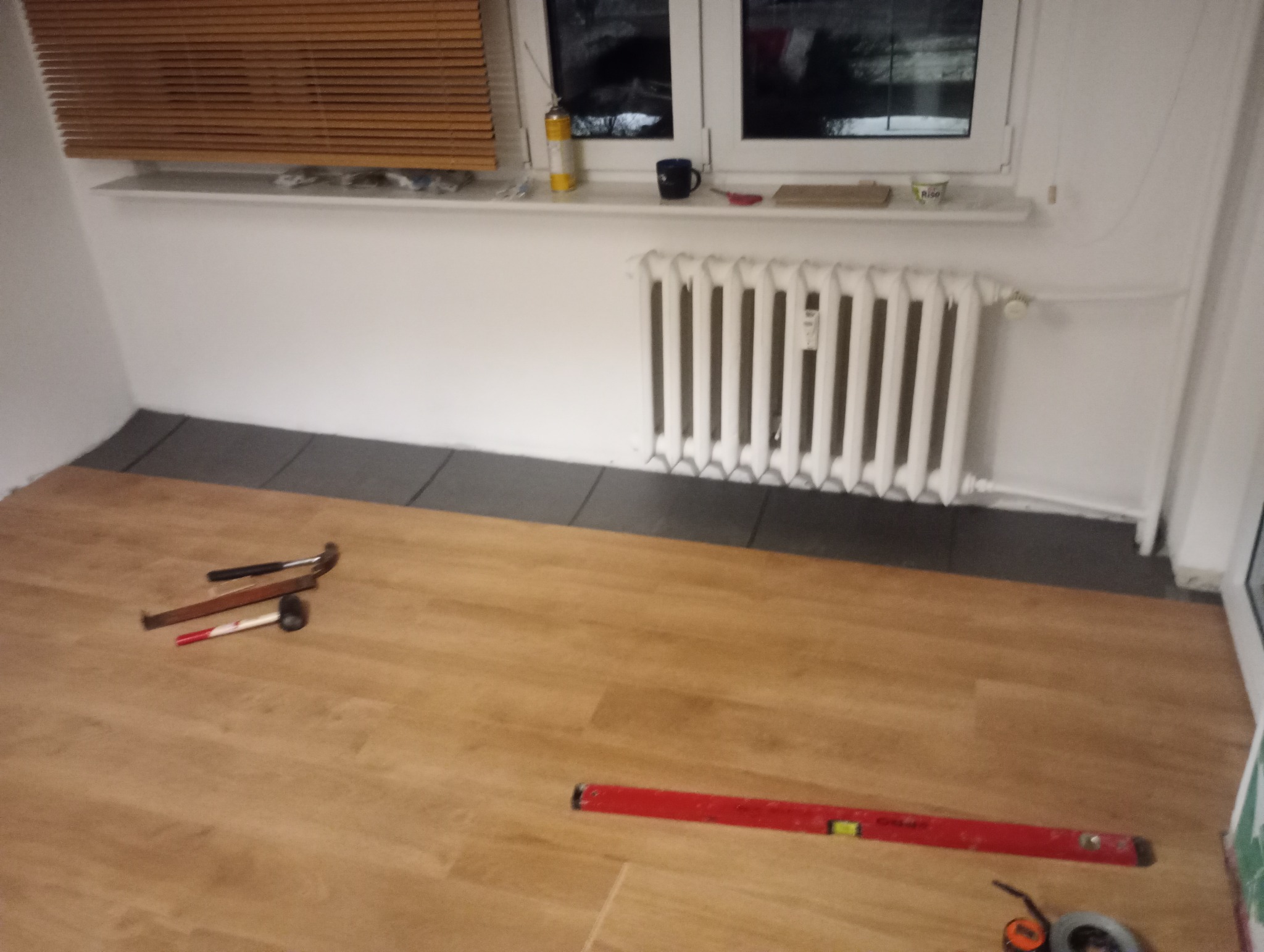 Fragment pokoju z ułożonymi panelami podłogowymi w odcieniach brązu. Widoczne narzędzia: młotek, poziomica. Przy ścianie grzejnik i podkład pod panele.