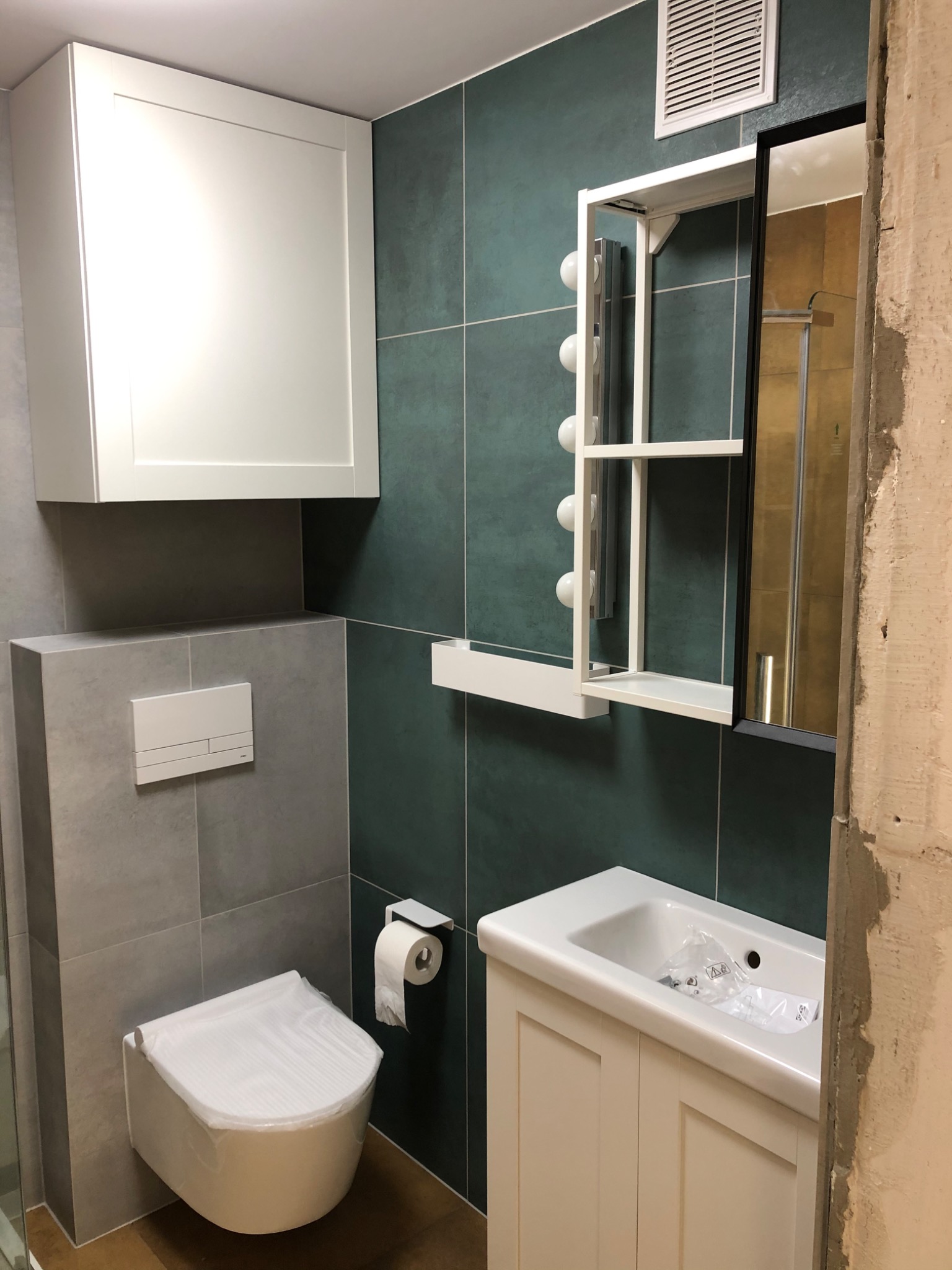 Nowoczesna łazienka w trakcie wykańczania: wisząca toaleta z zabezpieczeniem, umywalka z szafką, lustro z oświetleniem i zielone płytki na ścianach.