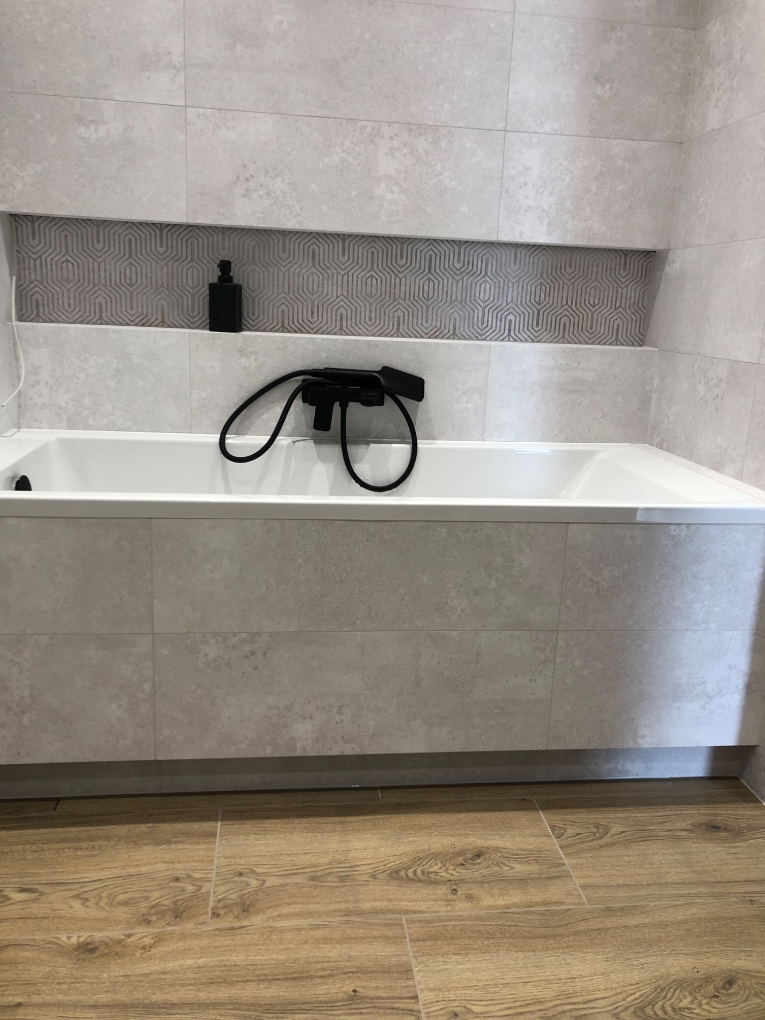 Nowoczesna łazienka z wanną obłożoną szarymi płytkami, czarna bateria wannowa i geometryczny wzór w niszy ściennej, uzupełniona butelką z dozownikiem i drewnopodobną podłogą.