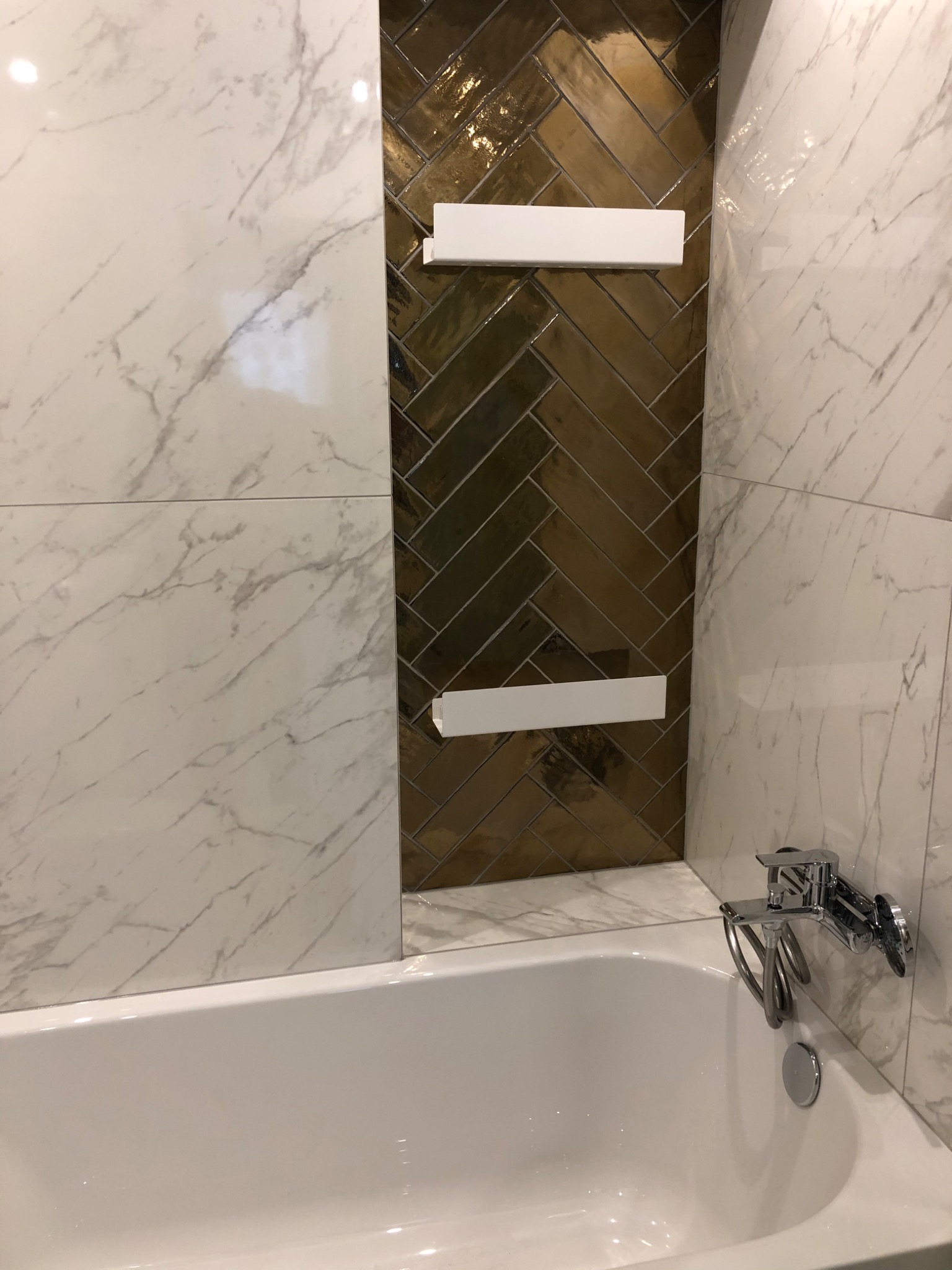 Wykończona łazienka z wanną, baterią wannową i złotymi płytkami ułożonymi w jodełkę, uzupełniona marmurowymi płytkami i dwoma białymi półkami.