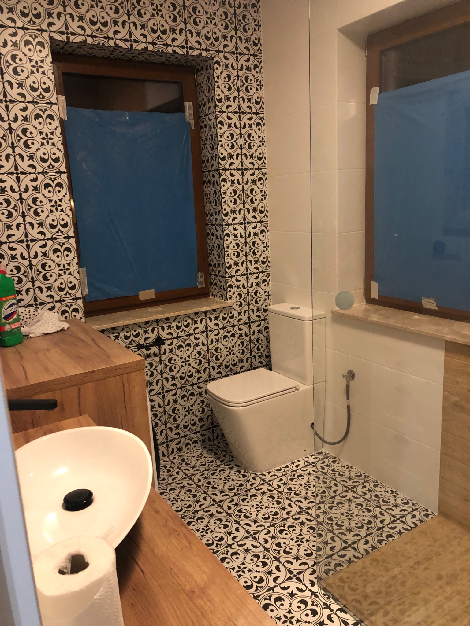 Ukończona łazienka z białą ceramiką, wzorzystymi czarno-białymi płytkami na ścianach i podłodze, drewnianą szafką pod umywalką i oknami zabezpieczonymi folią malarską.