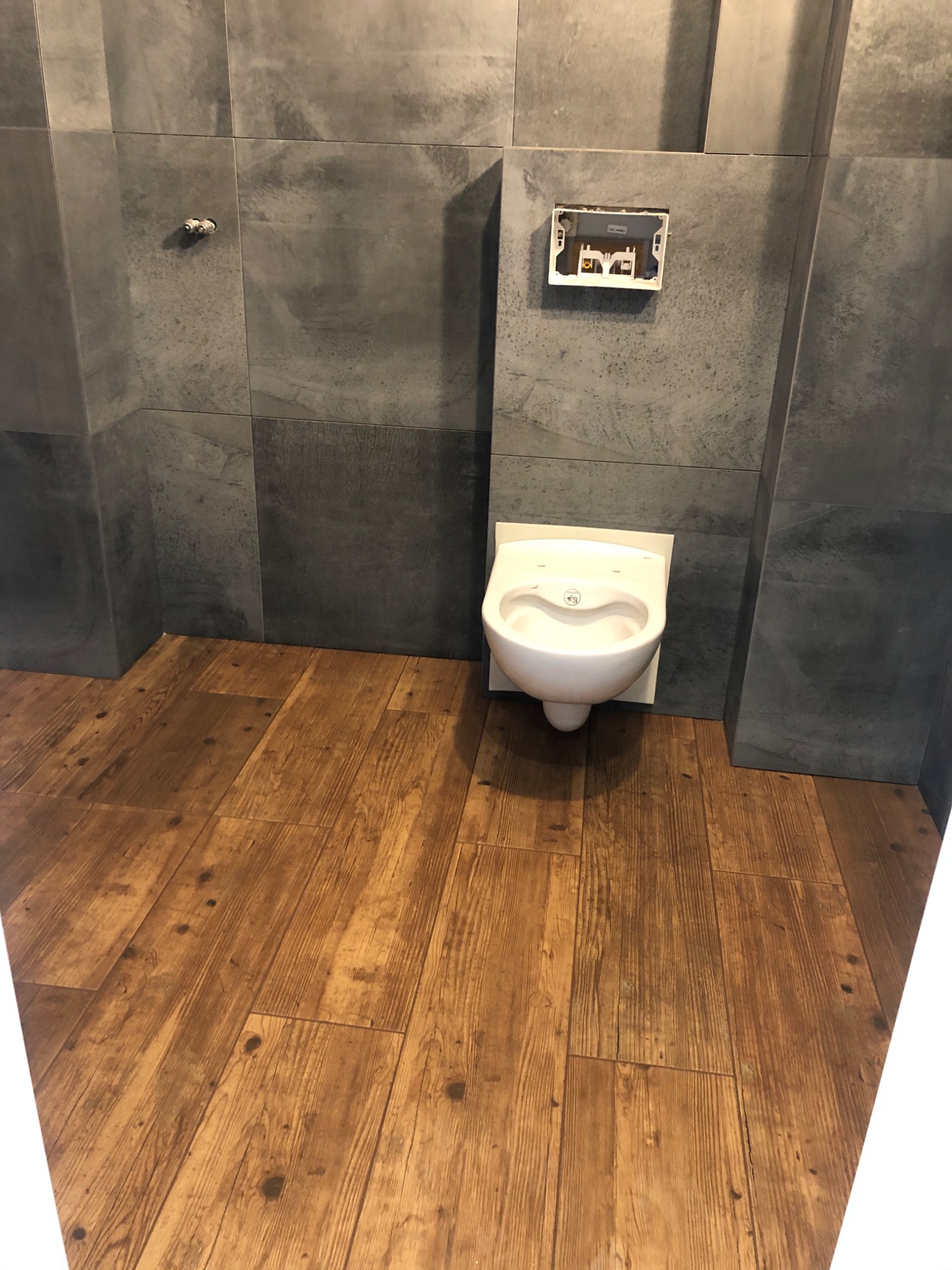 Wisząca miska WC z zamontowanym stelażem podtynkowym na tle szarych płytek i drewnopodobnej podłogi w trakcie montażu.