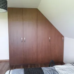 Brązowa szafa wnękowa z czterema drzwiami z podłużnymi, srebrnymi uchwytami, dopasowana do skośnego sufitu na poddaszu, widoczna drabinka u dołu.