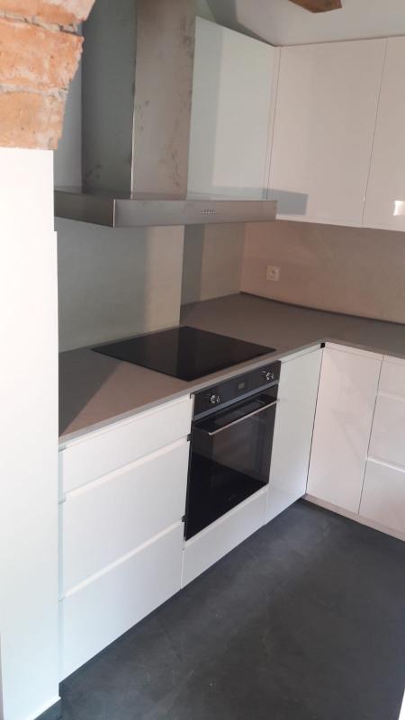 Nowoczesna, minimalistyczna zabudowa kuchenna z białymi, lakierowanymi frontami, szarym blatem i czarną płytą indukcyjną oraz piekarnikiem w zabudowie, widoczny fragment ceglanej ściany.