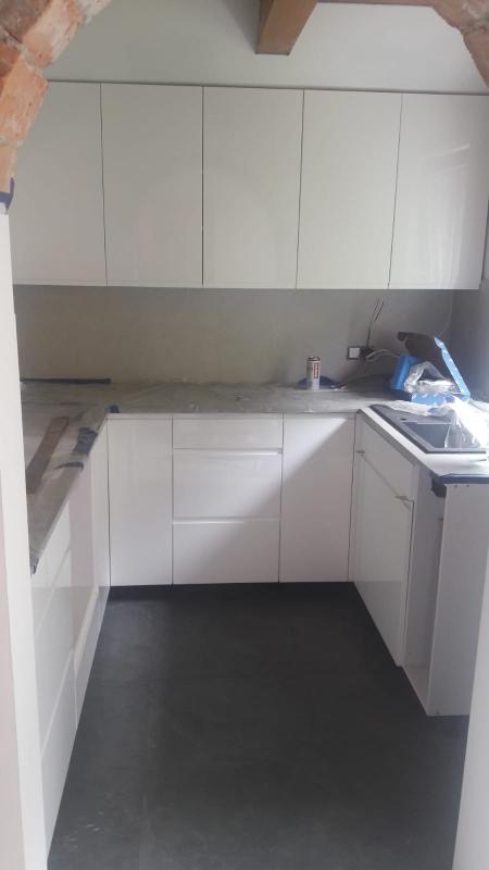 Nowoczesna kuchnia z białymi, lakierowanymi frontami szafek, szarym blatem i ciemną podłogą w trakcie montażu, widoczne narzędzia i materiały budowlane.