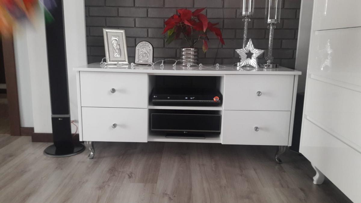 Biała szafka RTV z szufladami i srebrnymi nóżkami, na blacie dekoracje świąteczne i sprzęt elektroniczny, tło z cegły.