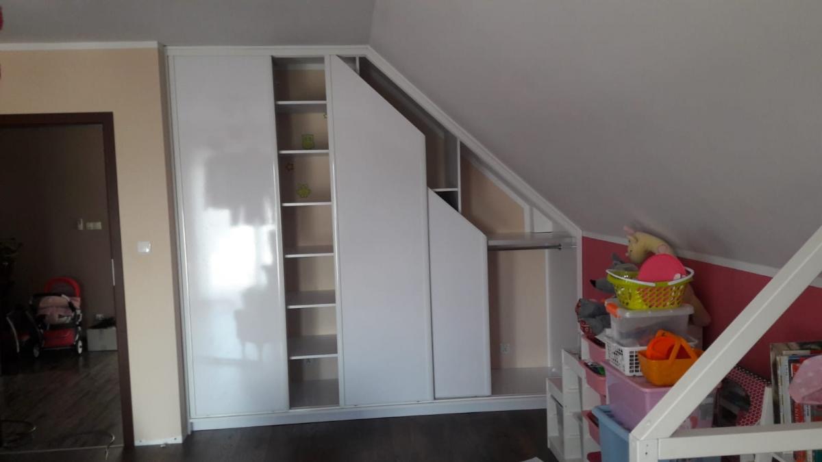 Biała szafa wnękowa z przesuwnymi drzwiami, dopasowana do skosu poddasza, z widocznymi półkami i przestrzenią na wieszaki, w pokoju dziecięcym z zabawkami w tle.