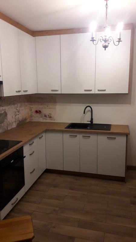 Kuchnia z białymi szafkami, drewnianym blatem i podłogą, czarnym zlewem i baterią, oraz wiszącym żyrandolem, ściana częściowo wykończona, częściowo z widoczną strukturą cegły.