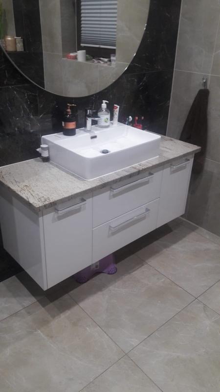 Biała szafka pod umywalkę z blatem imitującym kamień, kwadratowa umywalka nablatowa, duże owalne lustro, szare płytki na ścianie i podłodze, widoczne akcesoria łazienkowe.