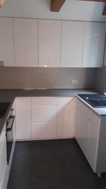 Nowoczesna kuchnia z białymi, lakierowanymi frontami szafek, szarym blatem i ciemną podłogą, widoczny fragment płyty grzewczej i zlewu.