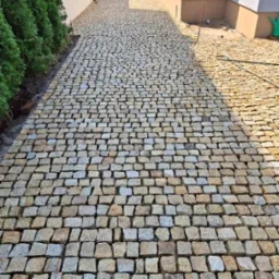 Ułożona droga z kostki brukowej w odcieniach szarości i żółci prowadząca do domu, widok z perspektywy osoby stojącej na początku drogi, z zielonymi krzewami po lewej stronie i domem po prawej stronie.